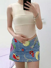 Person wearing a beige sleeveless top and a colorful checkered skirt with fruit designs.